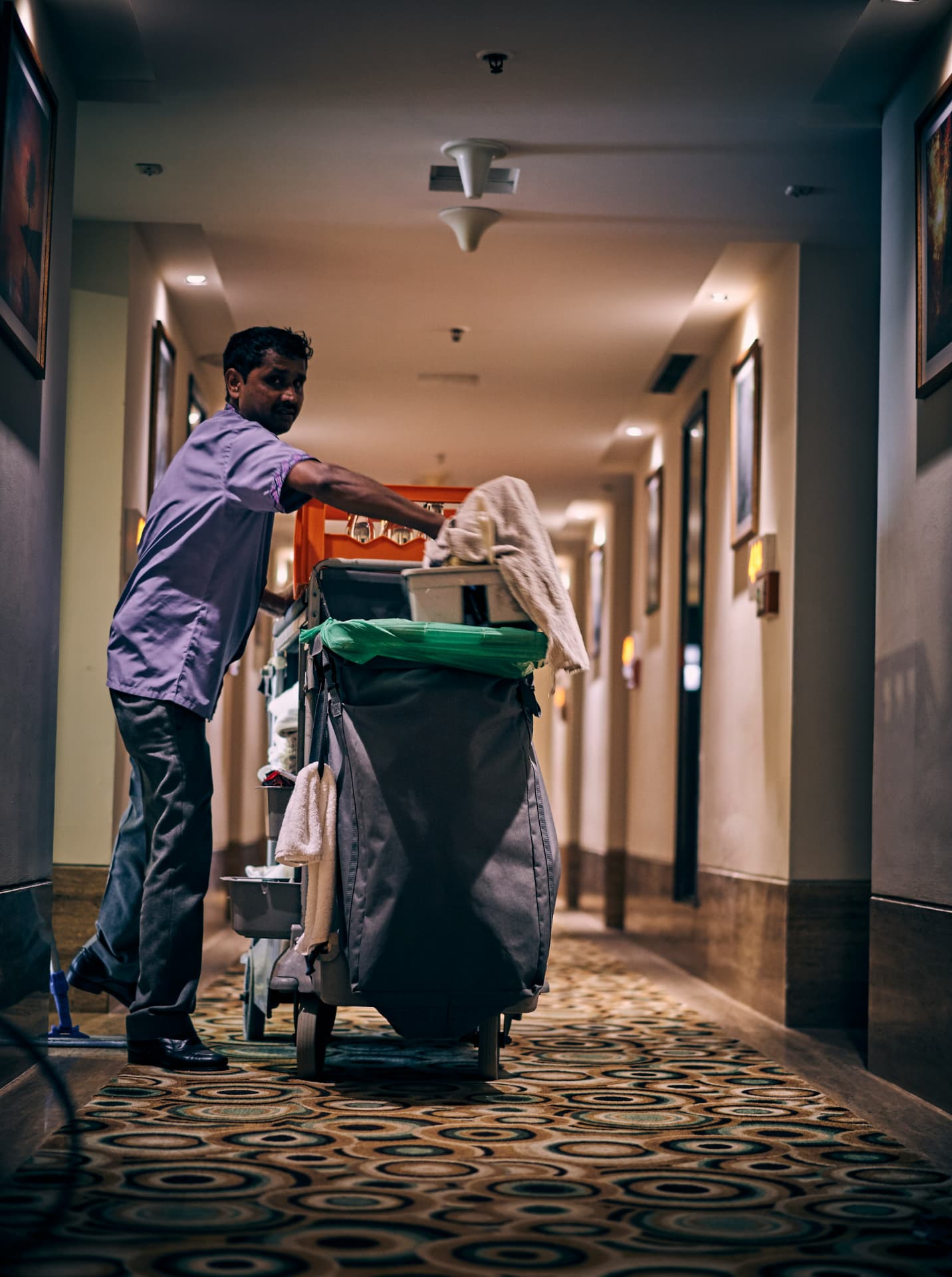 A housekeeper pushing a cleaning cart along a dim hostel corridor